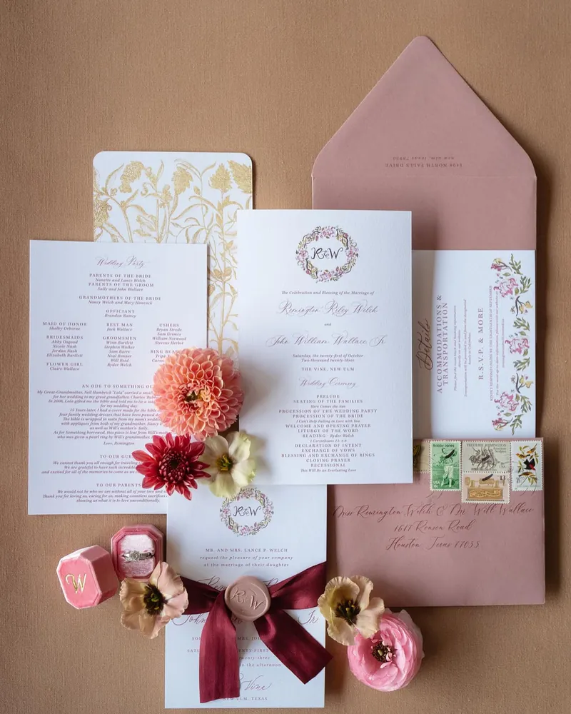 Blush wedding invitation suite with a floral wreath monogram, pink envelope, vintage stamps, and a burgundy silk ribbon