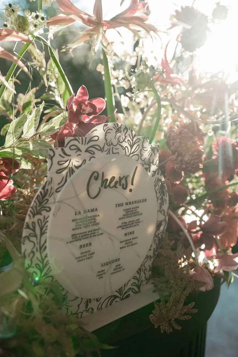 Clear acrylic "Cheers!" bar menu with black lettering set among lush garden florals